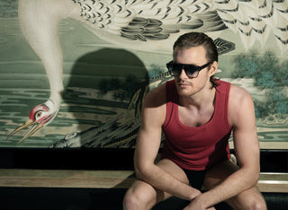 John VanBeber in red tank top wearing L'Ecurie Paris sunglasses before a crane mural, Fashion For Men editorial